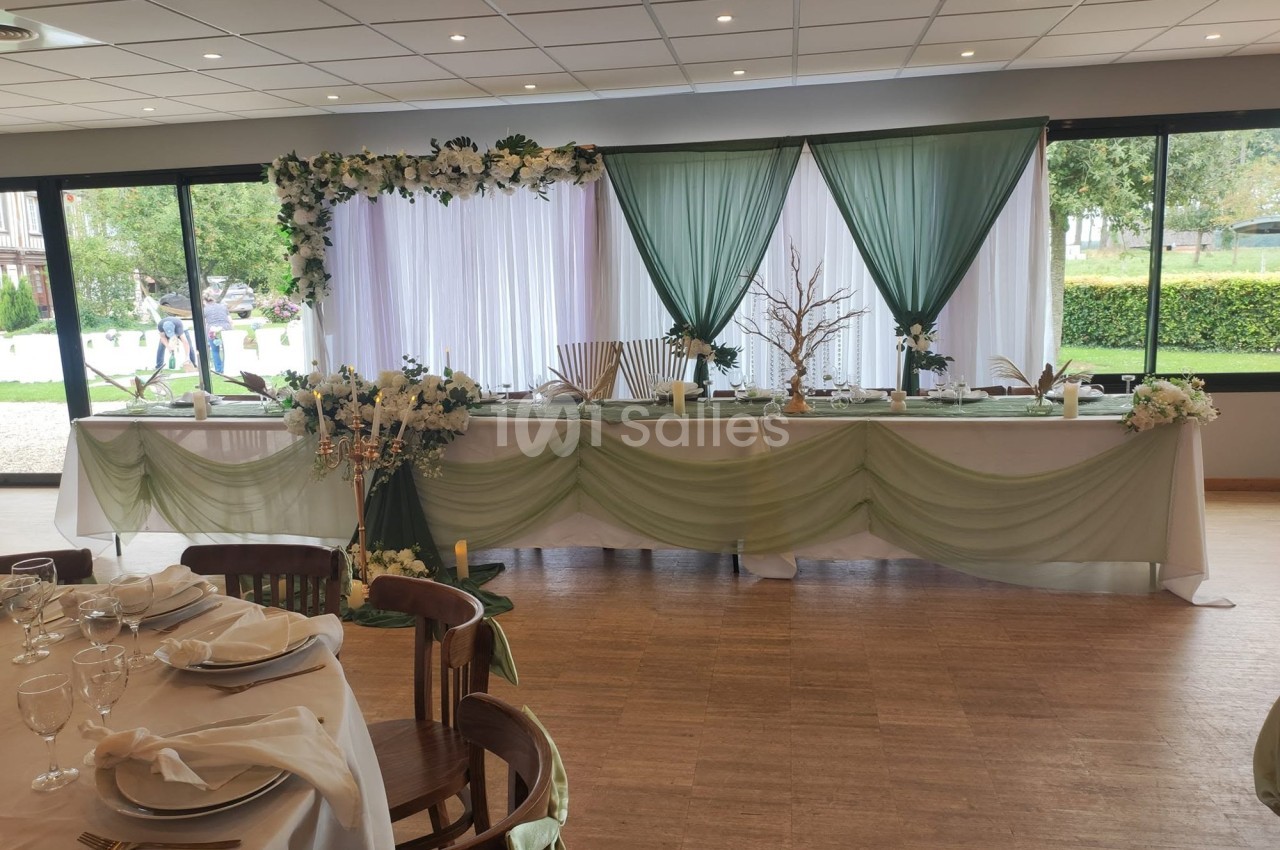 Salle de réception dressée avec une table d'honneur drapée de voile vert sauge, ornée de fleurs blanches, de bougies et d'un arbre décoratif, baies vitrées donnant sur un jardin.
