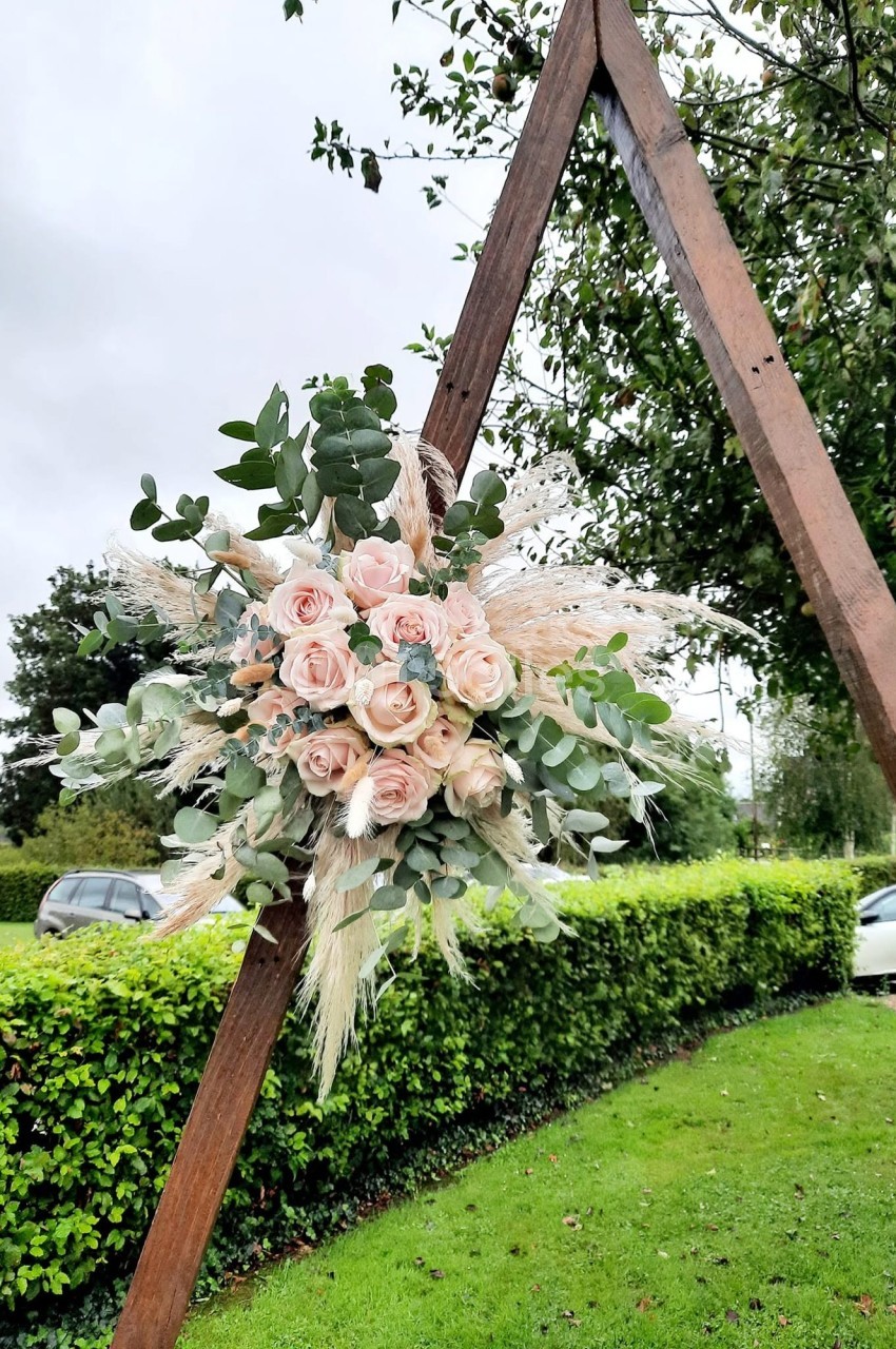 Arche triangulaire en bois foncé ornée d'un bouquet de roses rose pâle, d'eucalyptus et de graminées séchées, sur une pelouse bordée de haies.
