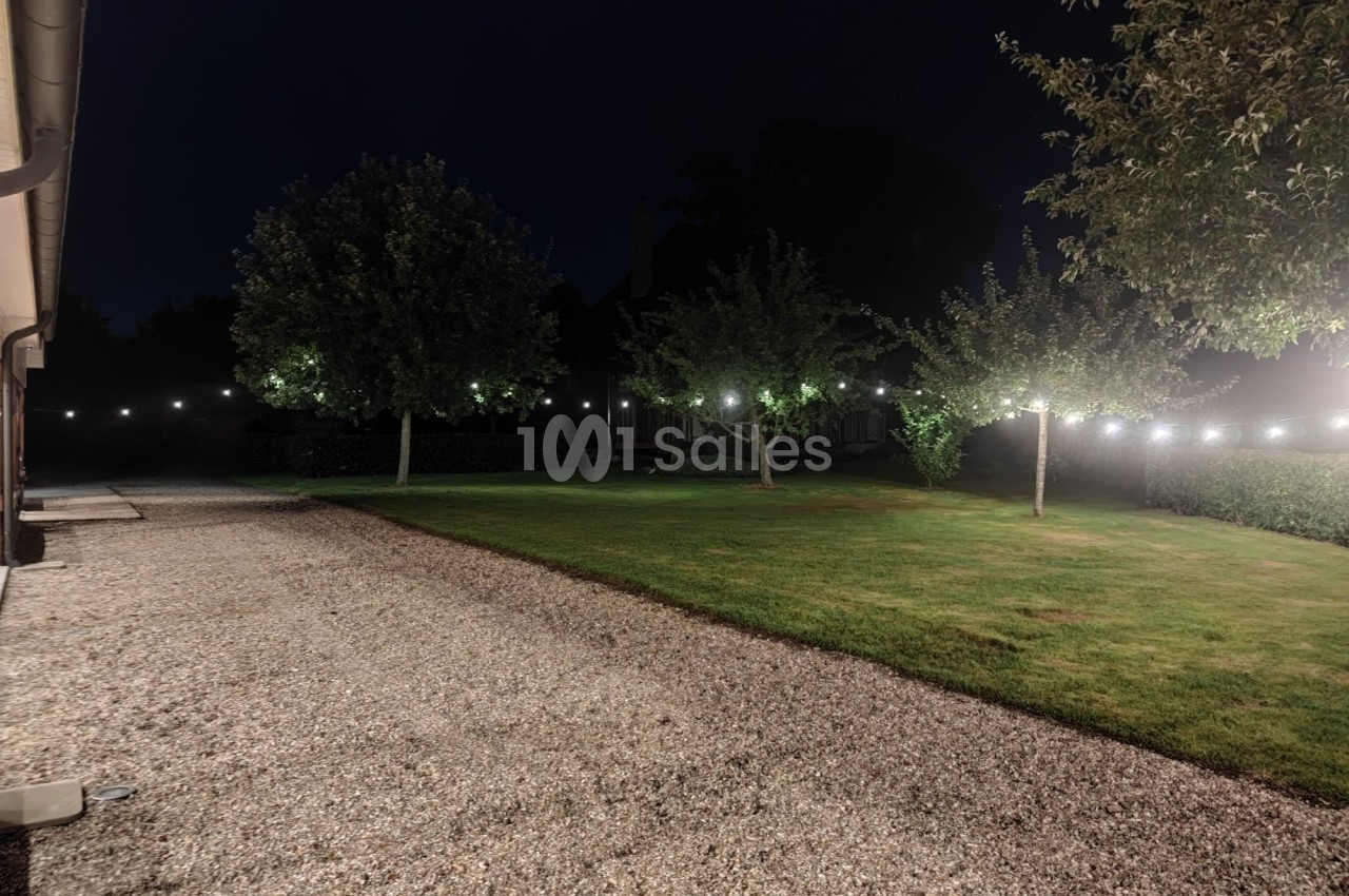 Jardin engazonné de nuit, éclairé par des guirlandes lumineuses suspendues entre les arbres, bordé d'une haie et d'une allée de gravier.