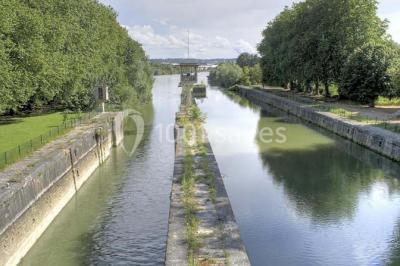 Canal bordé d'arbres avec une écluse centrale et un paysage verdoyant sous un ciel partiellement nuageux.