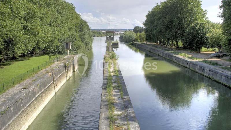 Canal bordé d'arbres avec une écluse centrale et un paysage verdoyant sous un ciel partiellement nuageux.