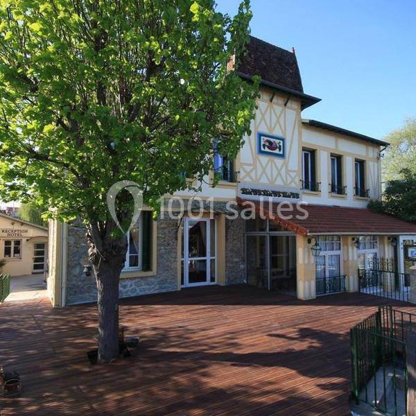 Façade d'un bâtiment à colombages avec terrasse en bois, entouré d'arbres sous un ciel bleu.
