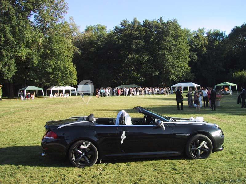 Voiture décapotable noire décorée pour un mariage, garée sur une pelouse avec des invités et des tentes en arrière-plan.