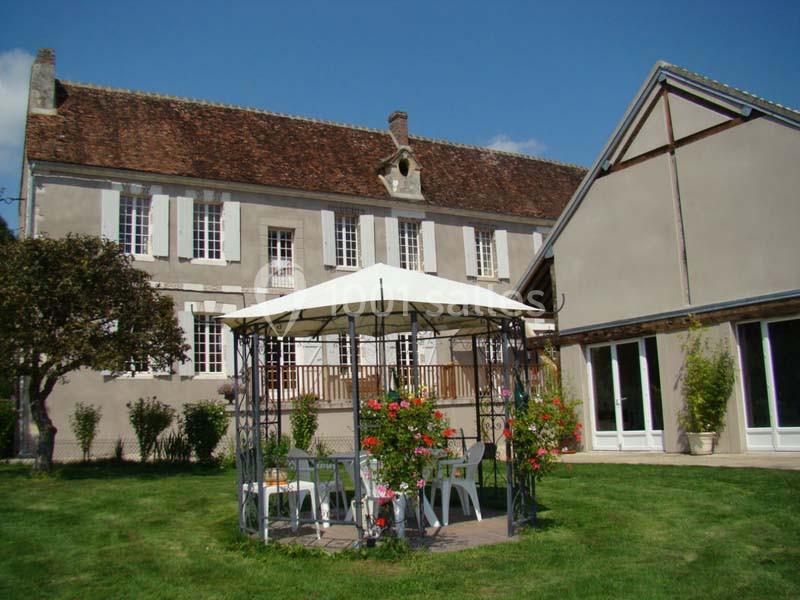 Façade d'une maison ancienne avec un jardin, une tonnelle blanche et des chaises disposées à l'ombre.