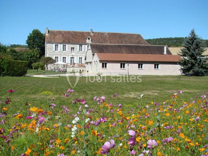 Grande maison en pierre entourée d'un jardin fleuri avec des champs et des collines en arrière-plan.