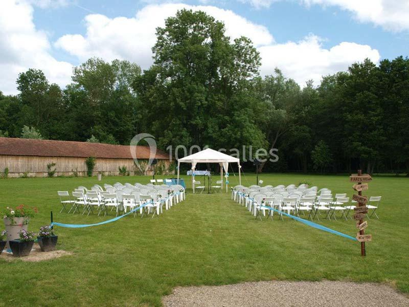 Chaises blanches disposées en extérieur devant une tonnelle sur une pelouse, entourées d'arbres et d'un bâtiment en bois.
