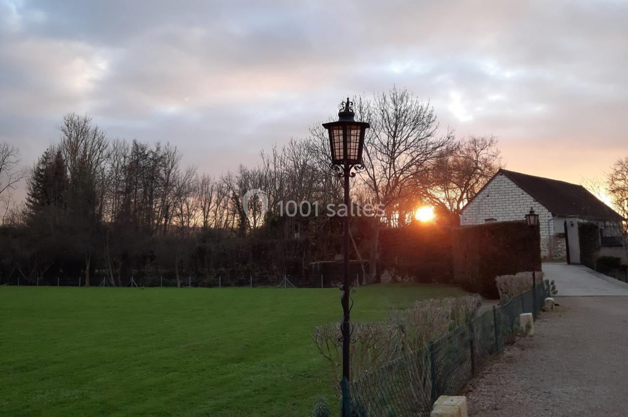 Un lampadaire en fer forgé près d'une allée, avec un coucher de soleil derrière des arbres et une maison en pierre.