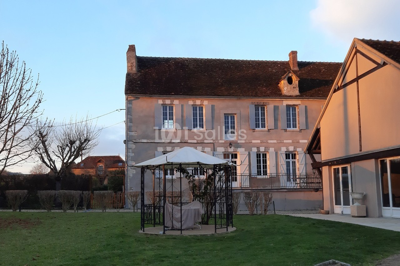 Maison ancienne en pierre avec volets blancs, jardin arboré et tonnelle au centre, sous un ciel partiellement dégagé.