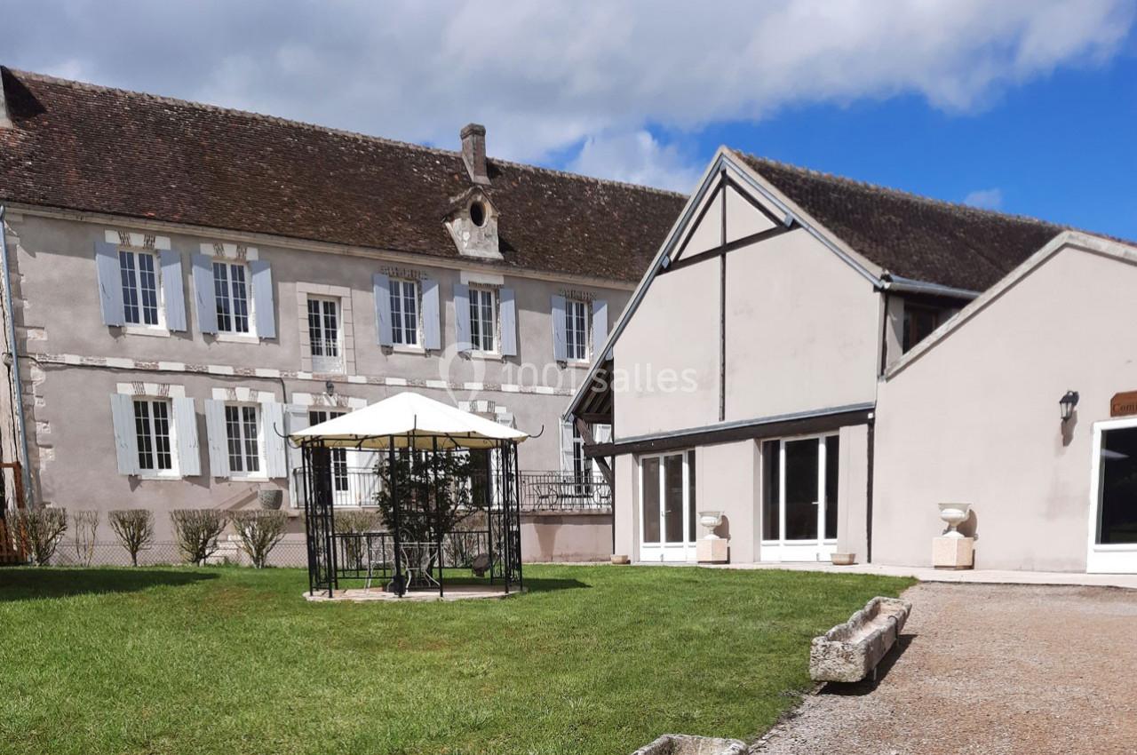 Cour extérieure avec pelouse, tonnelle en métal et bâtiments anciens aux façades claires sous un ciel partiellement nuageux.