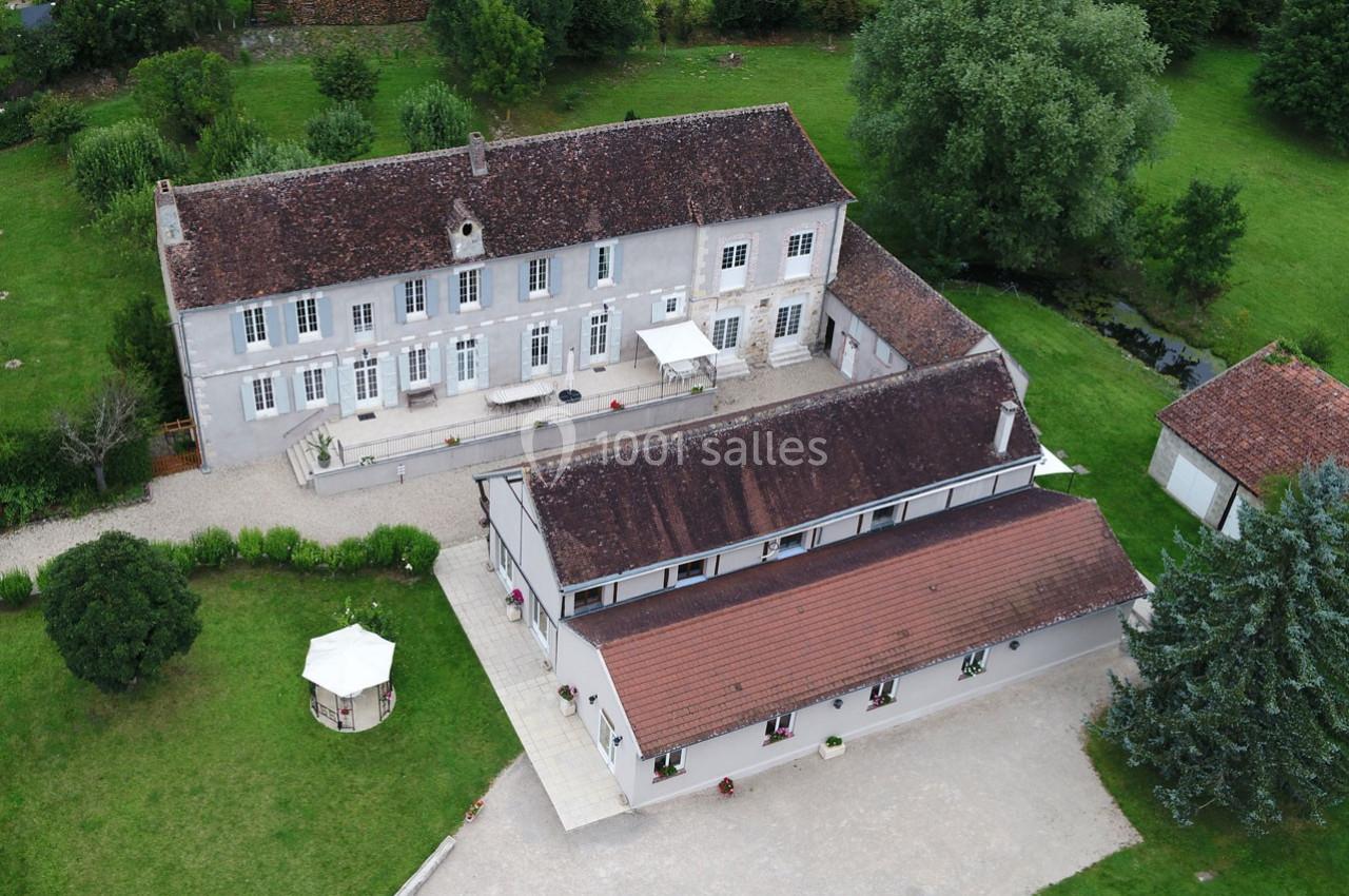 Vue aérienne d'un grand bâtiment ancien avec toiture en tuiles, entouré de jardins et d'arbres verdoyants.
