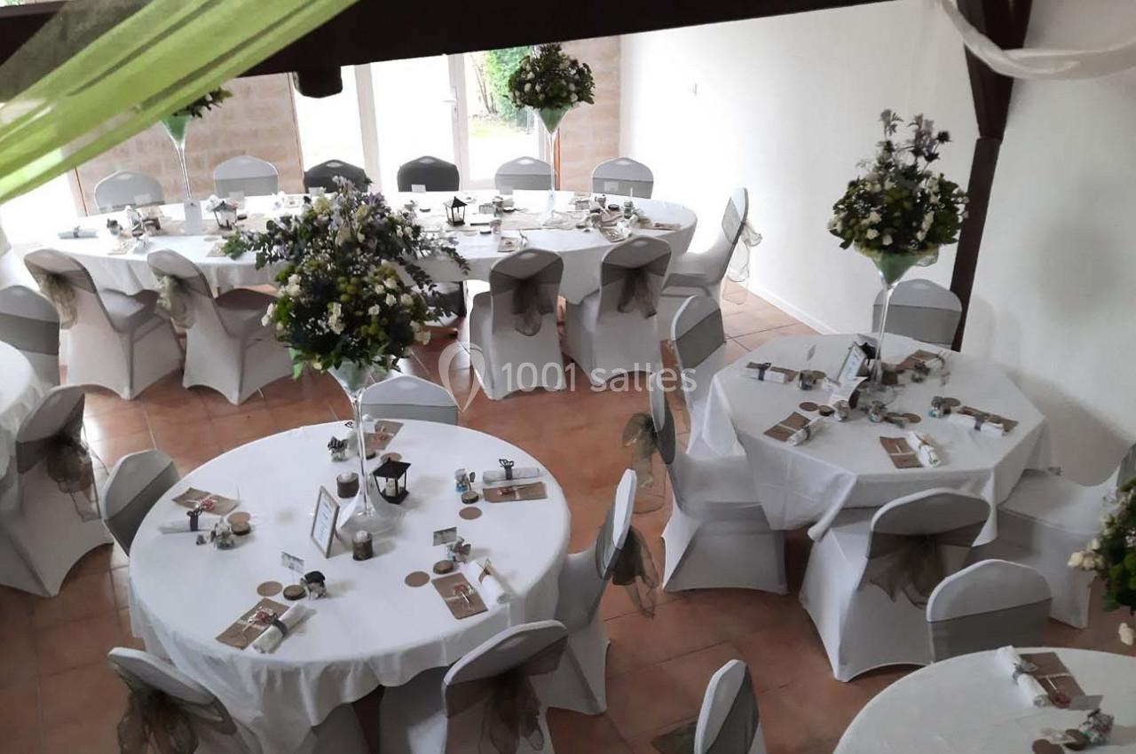 Salle décorée pour un événement avec des tables rondes dressées, nappes blanches et centres de table fleuris.