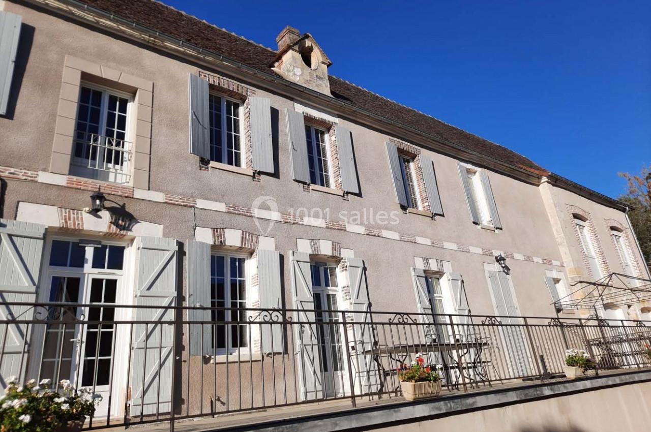 Façade d'une maison ancienne avec volets blancs, terrasse en métal et ciel bleu en arrière-plan.
