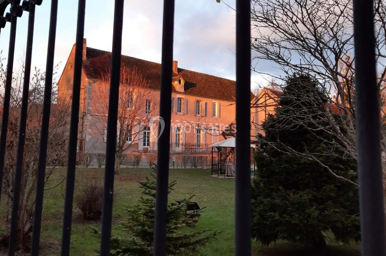 Manoir en pierre éclairé par le soleil couchant, vu à travers une grille, entouré d'arbres et d'un jardin.