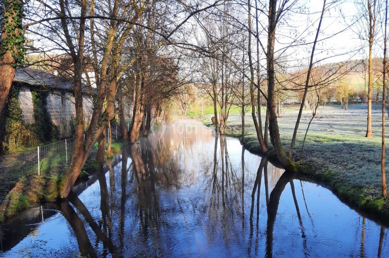 Rivière calme bordée d'arbres dénudés en hiver, avec des champs givrés et un bâtiment en pierre à gauche.