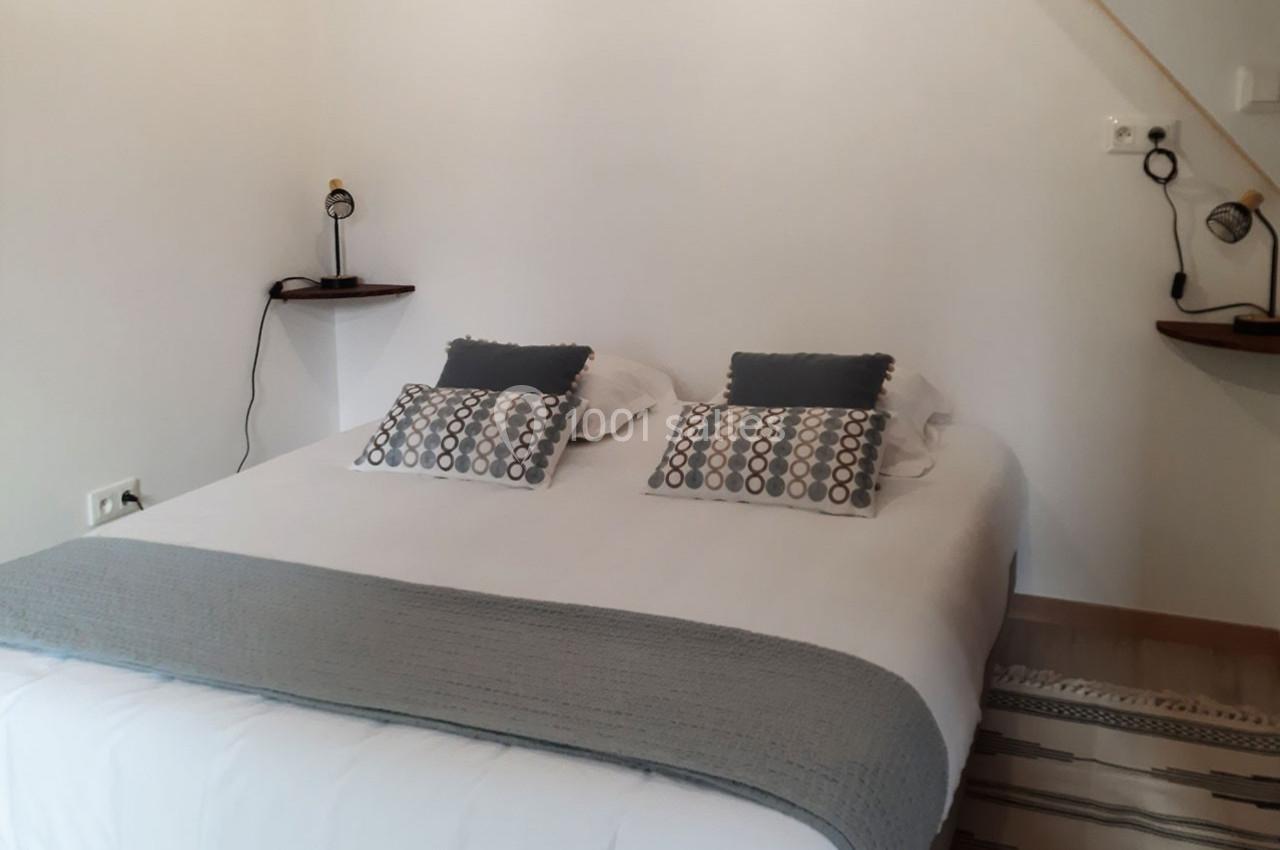 Lit double avec draps blancs, couverture grise, coussins décoratifs et lampes de chevet fixées au mur.