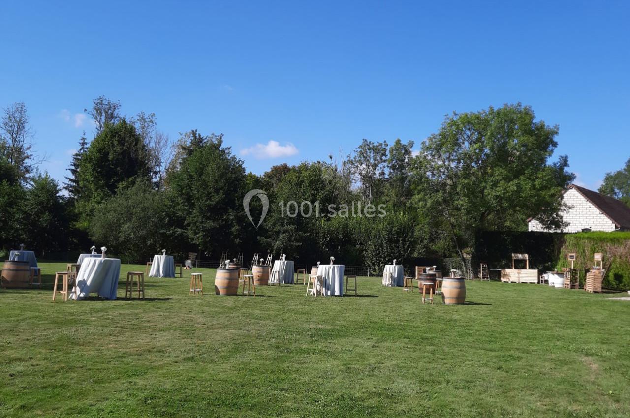 Tables hautes et tonneaux disposés sur une pelouse verdoyante, entourés d'arbres sous un ciel dégagé.