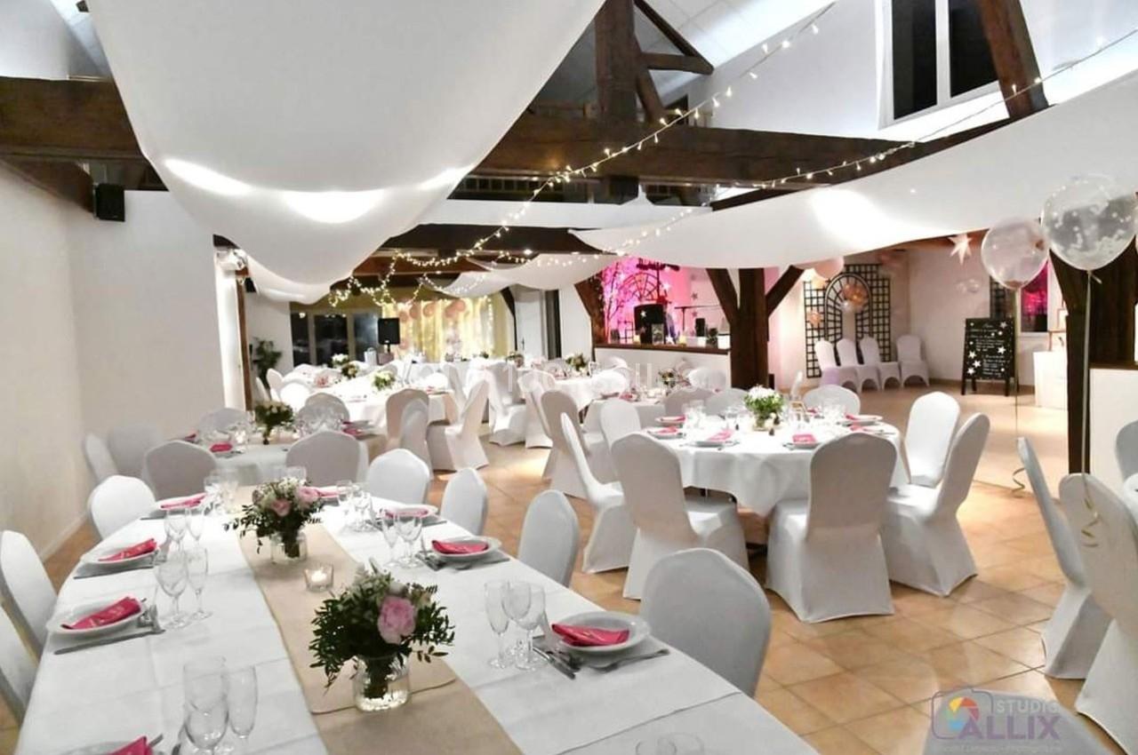 Salle de réception décorée avec des tables dressées, nappes blanches, chaises houssées et guirlandes lumineuses suspendues.