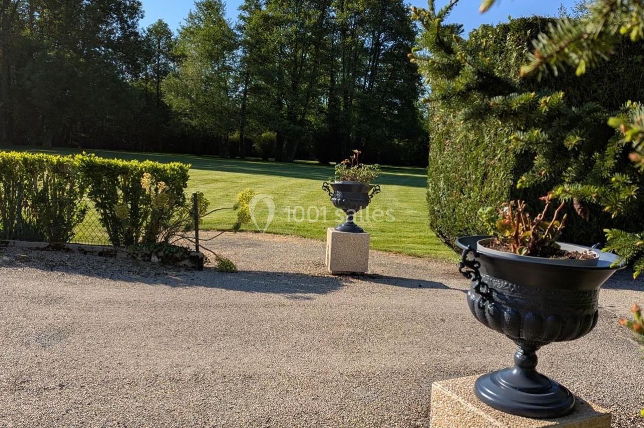 Allée bordée de jardinières en métal noir, donnant sur une pelouse entourée d'arbres et de haies.