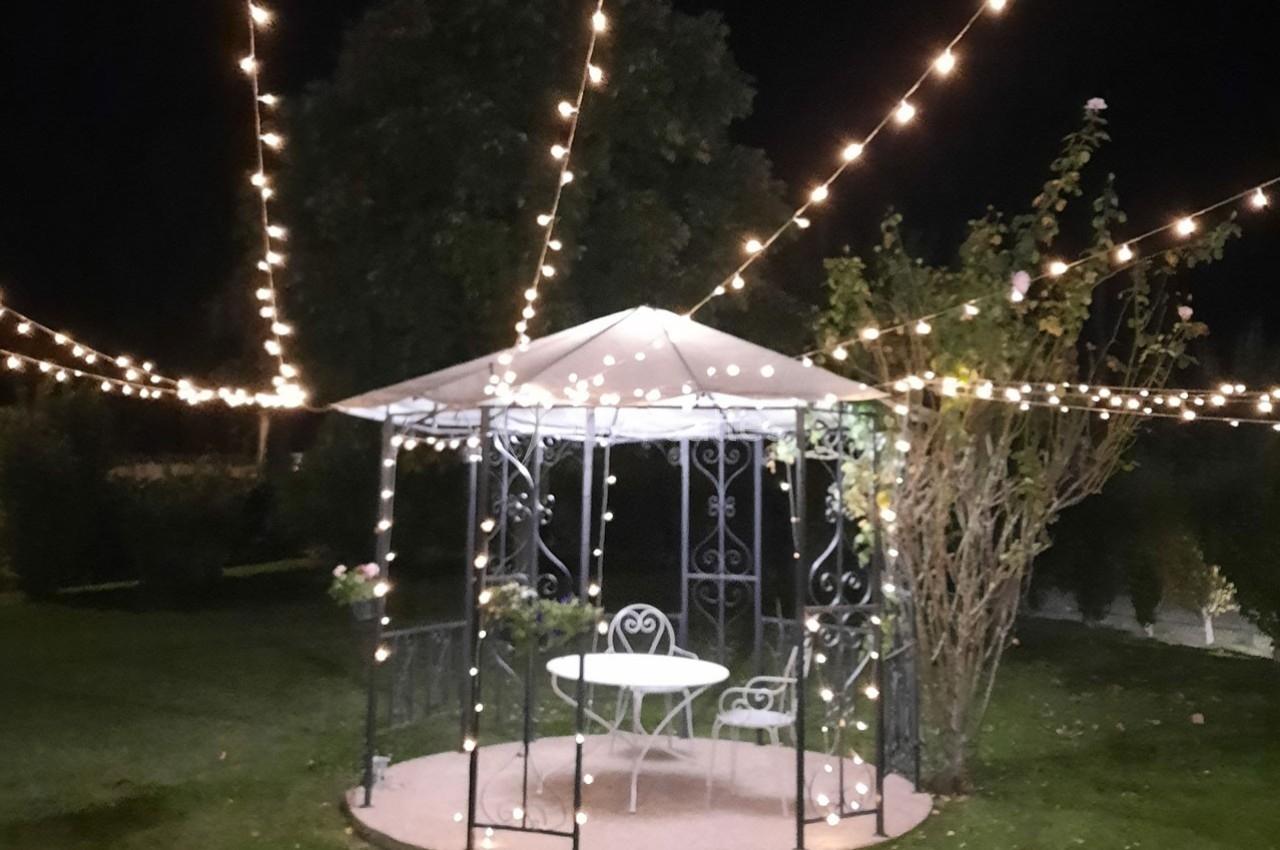 Tonnelle de jardin éclairée par des guirlandes lumineuses, avec une table et des chaises blanches, de nuit.