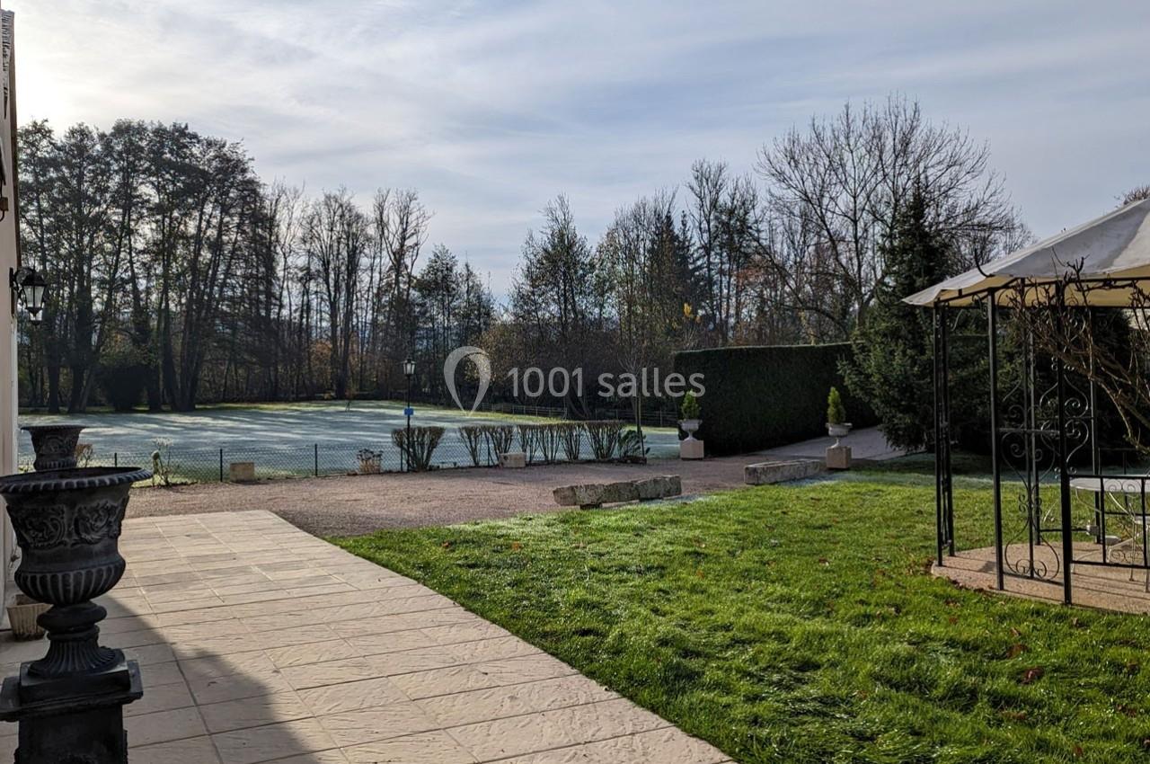 Vue d'un jardin avec pelouse, arbres en arrière-plan, terrasse pavée et pergola métallique sur la droite.