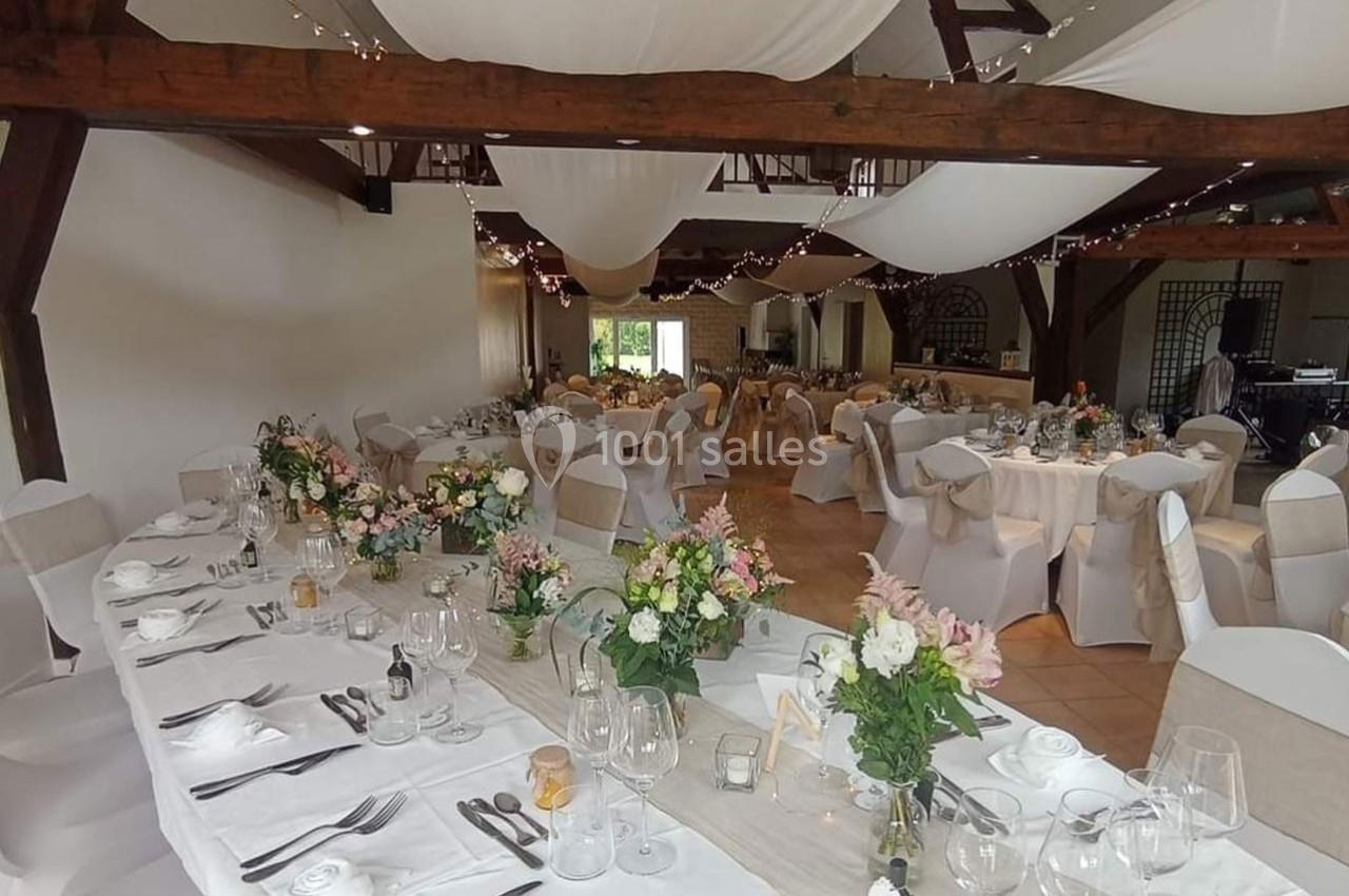 Salle de réception décorée pour un mariage avec tables dressées, nappes blanches, fleurs et chaises ornées de nœuds beiges.