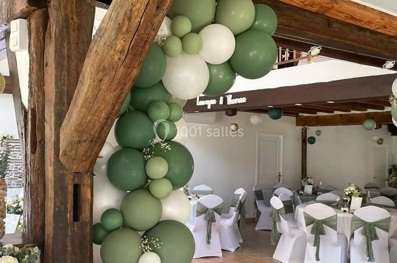 Décoration de salle avec arche de ballons verts et blancs, chaises habillées de nœuds verts et tables dressées.