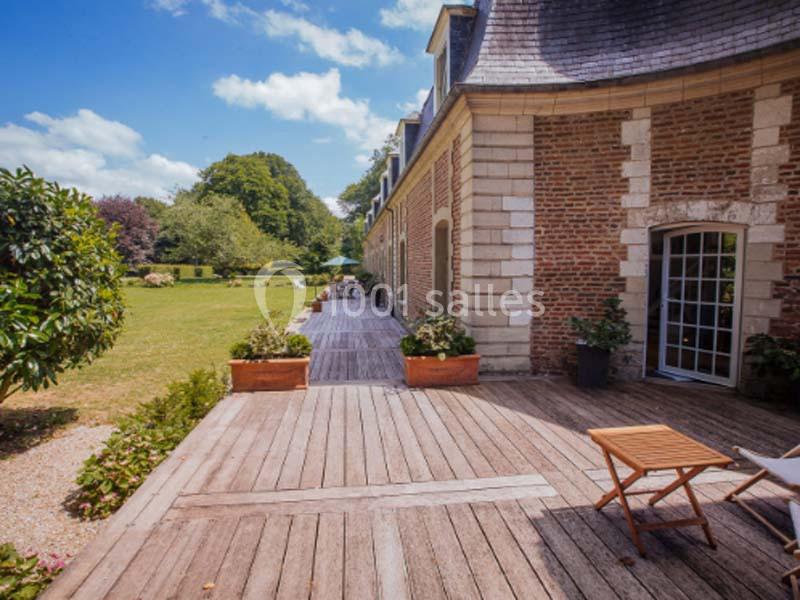 Terrasse en bois longeant un bâtiment en briques avec vue sur un jardin arboré sous un ciel bleu.
