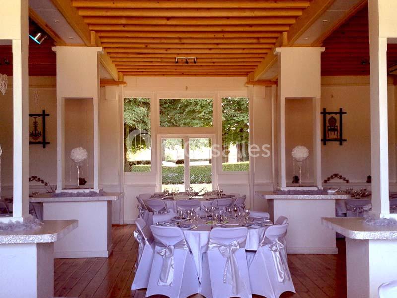 Salle de réception lumineuse avec tables dressées, chaises décorées de rubans et vue sur un jardin verdoyant.