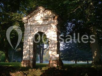 Vestige d'une petite arche en pierre entourée d'arbres dans un parc verdoyant éclairé par une lumière douce.