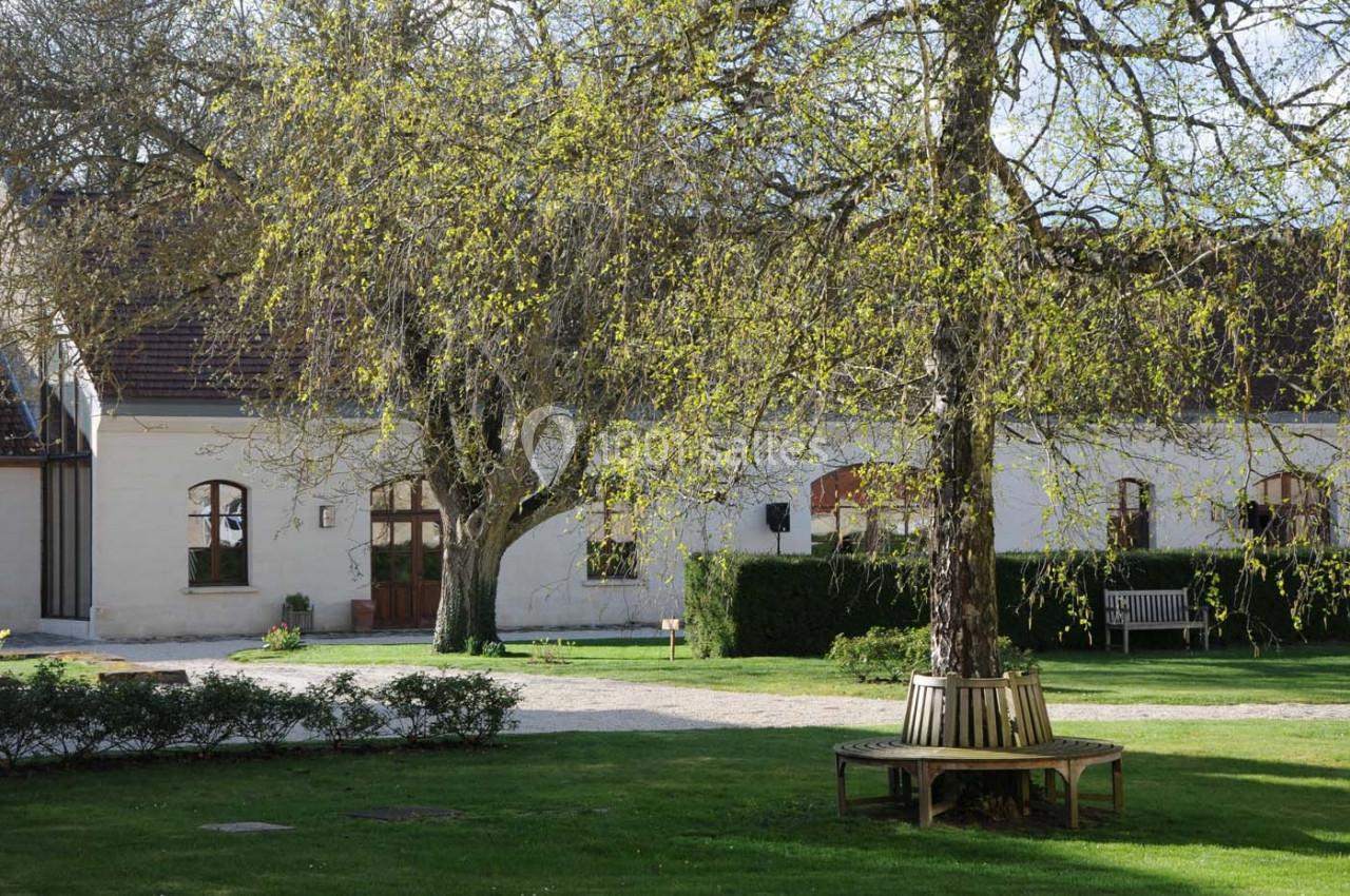 Cour verdoyante avec banc circulaire autour d’un arbre, devant un bâtiment aux murs clairs et fenêtres en arc.