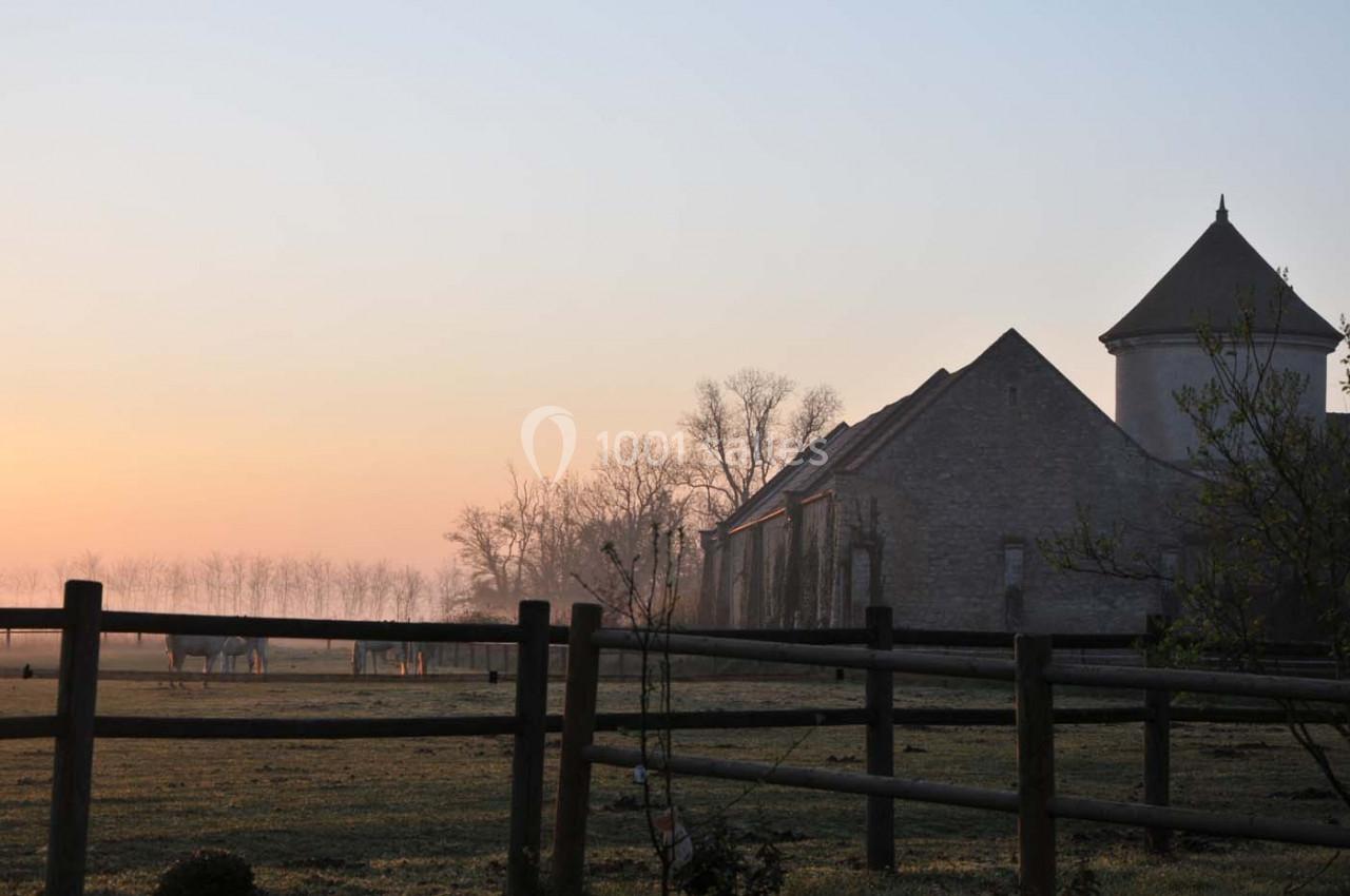 Vue d'un bâtiment en pierre avec une tour, entouré de champs et de chevaux au lever du soleil.