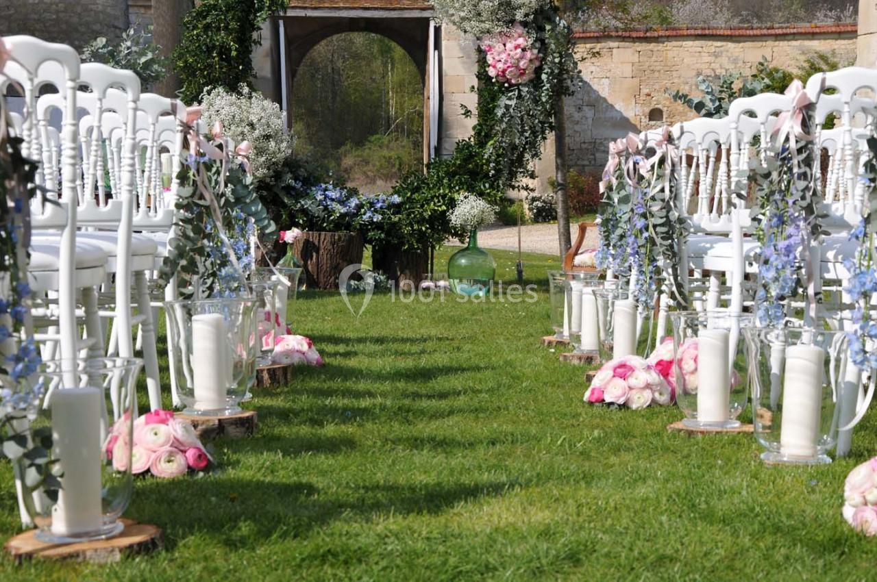 Allée centrale décorée de fleurs et bougies, bordée de chaises blanches, menant à une arche florale en extérieur.