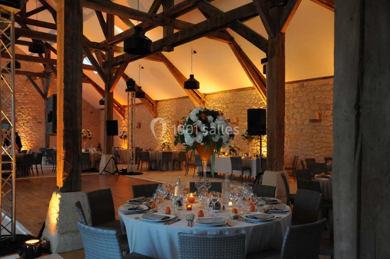 Salle de réception avec poutres apparentes, tables dressées, décorations florales et éclairage chaleureux.