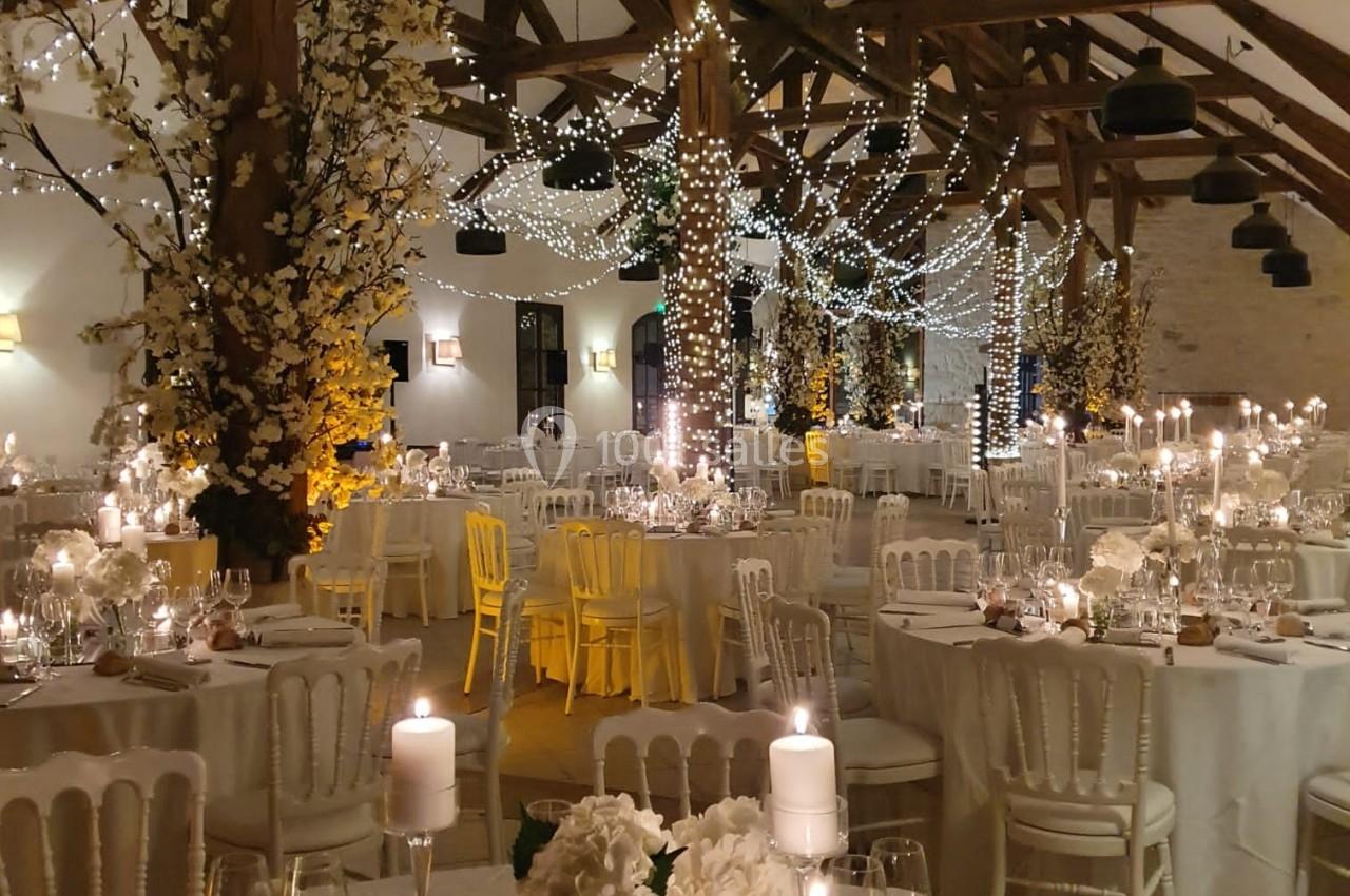 Salle de réception décorée pour un mariage, avec tables dressées, bougies, guirlandes lumineuses et arbres fleuris.