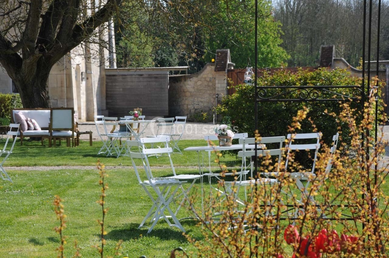 Chaises et tables blanches disposées sur une pelouse, entourées de verdure et de fleurs, dans un cadre extérieur calme.