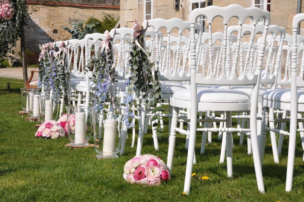 Chaises blanches alignées sur une pelouse, décorées de fleurs et de rubans pour une cérémonie en extérieur.