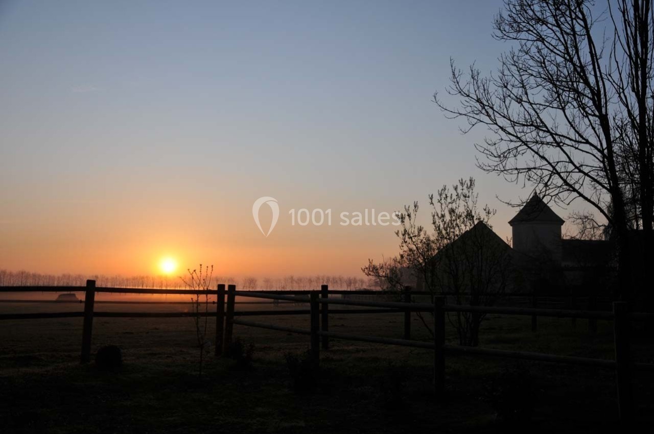 Lever de soleil sur un paysage rural avec une clôture en bois, des arbres nus et un bâtiment en arrière-plan.