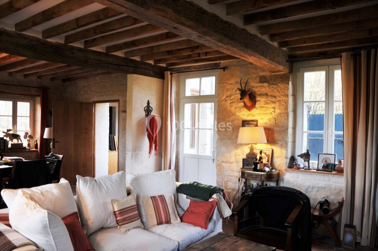 Salon chaleureux avec murs en pierre, poutres apparentes, canapé blanc, et lumière naturelle entrant par les fenêtres.