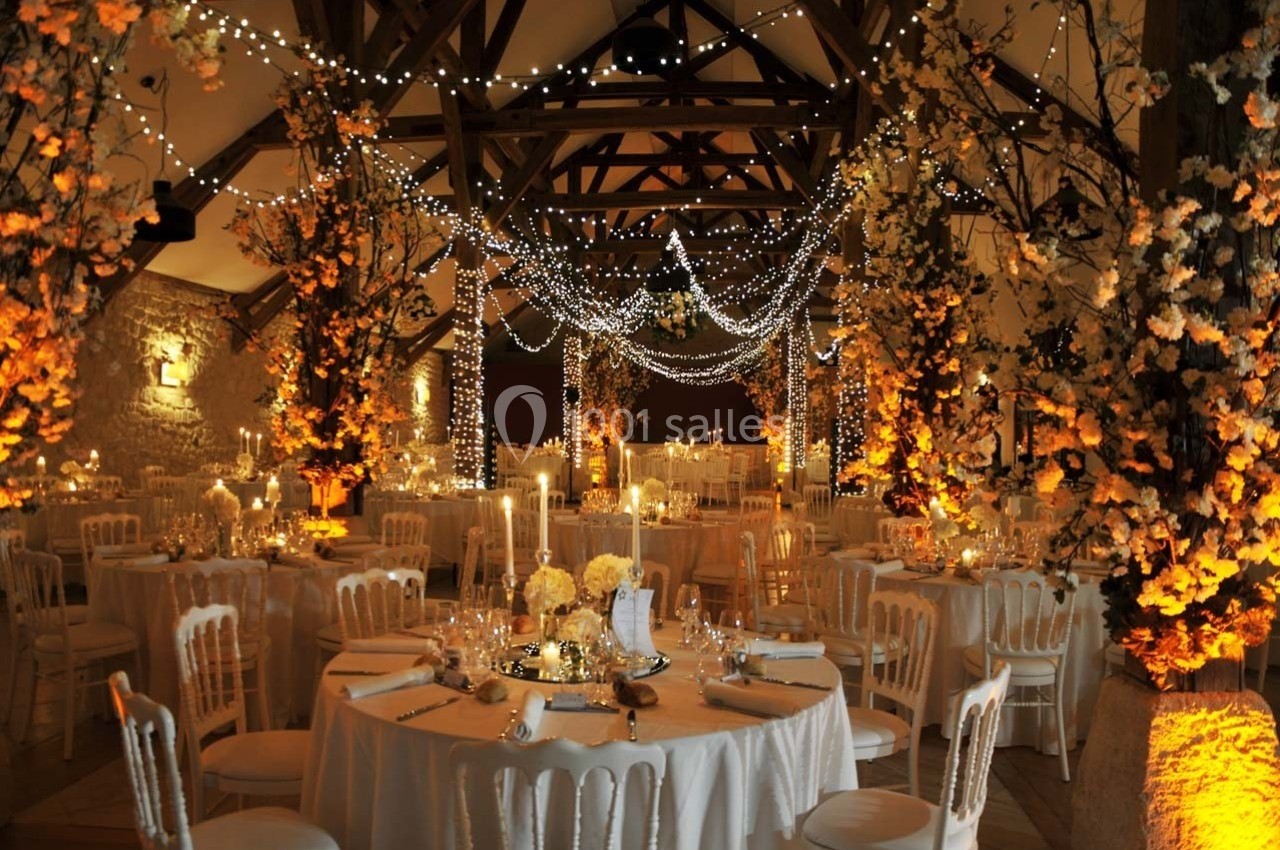 Salle de réception décorée pour un mariage, avec guirlandes lumineuses, fleurs et tables élégamment dressées.