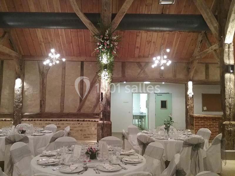 Salle de réception avec tables rondes dressées, nappes blanches, poutres apparentes et décoration florale suspendue.