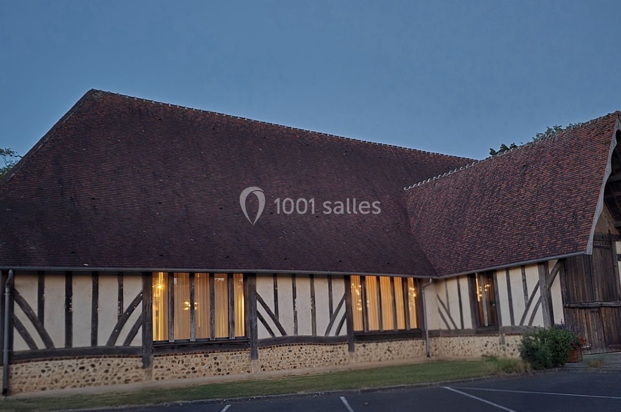 Bâtiment à colombages avec toit en pente, éclairé de l'intérieur, entouré de végétation et d'un parking.