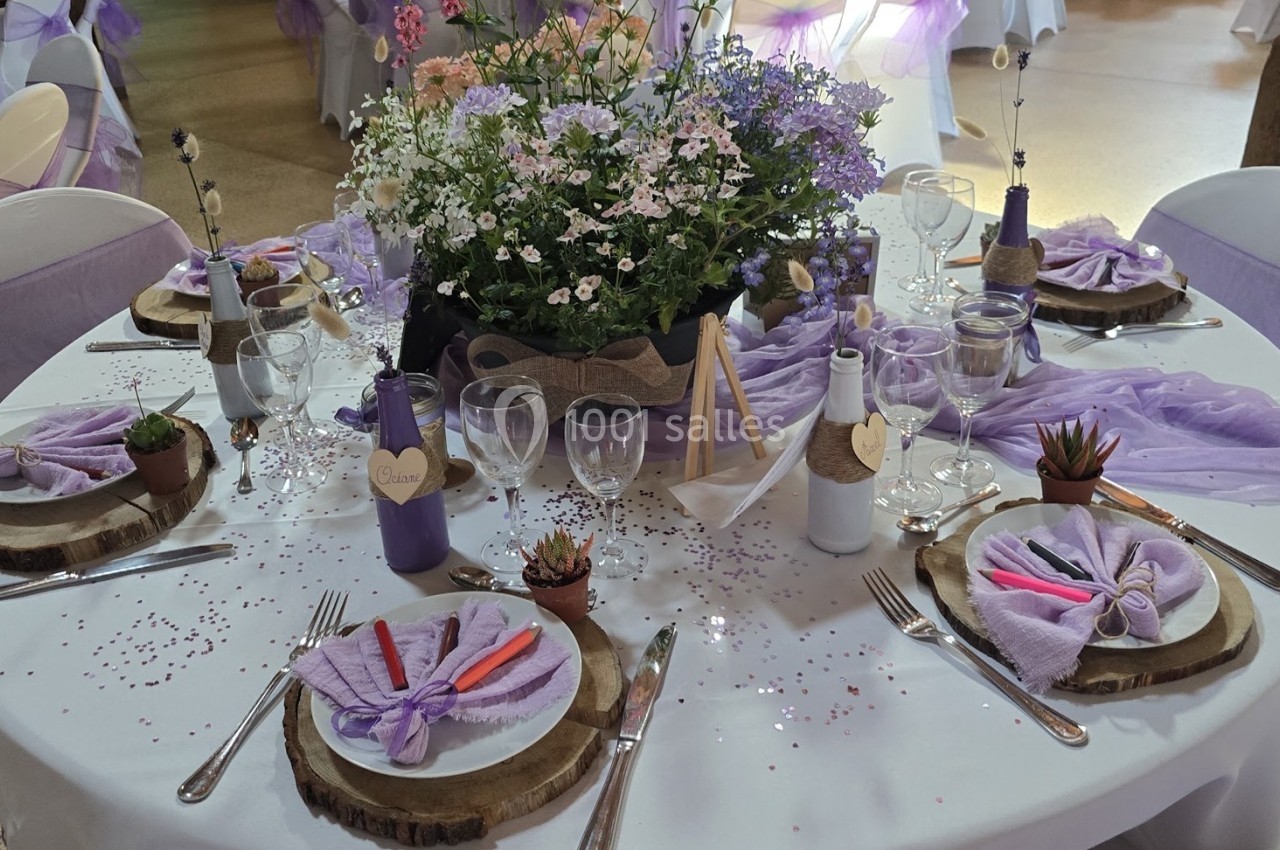 Table décorée pour un événement avec nappes violettes, fleurs, bougies, rondins de bois et couverts disposés.