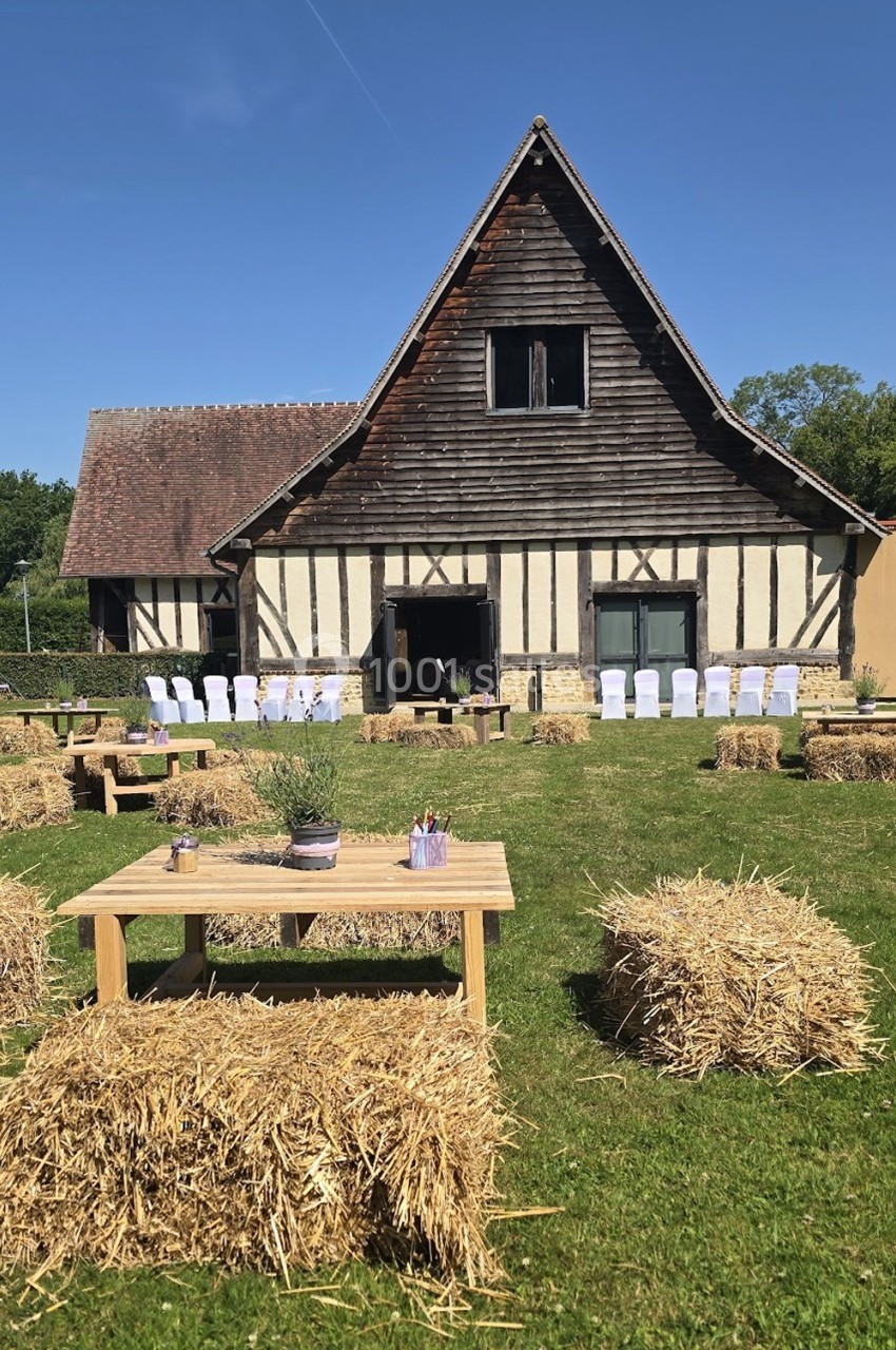Grange à colombages avec tables en bois et bottes de paille disposées sur une pelouse pour un événement en plein air.