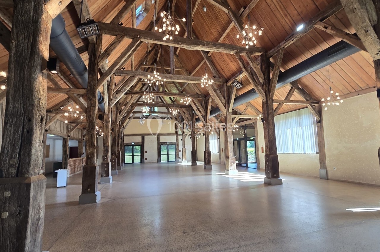 Grande salle avec charpente en bois apparente, sol en béton lisse, grandes fenêtres et éclairage suspendu.