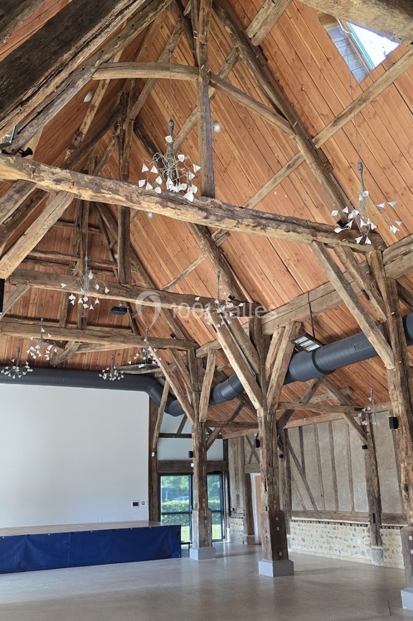 Charpente en bois apparente dans une grande salle lumineuse avec murs en pierre et écran blanc au fond.
