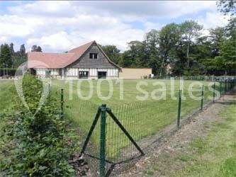 Bâtiment à colombages avec toit en pente, entouré d'une pelouse clôturée et d'arbres en arrière-plan.