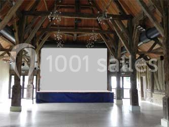 Salle avec charpente en bois apparente, grande scène surélevée et éclairage suspendu, baignée de lumière naturelle.