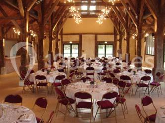 Salle de réception avec charpente en bois, tables rondes dressées et chaises rouges, éclairée par des lustres suspendus.