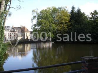 Vue d’un étang entouré d’arbres et de bâtiments anciens, avec une barrière au premier plan.