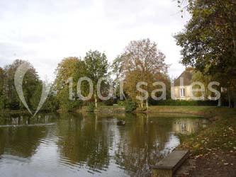 Étang calme entouré d'arbres en automne avec une maison en pierre au fond, sous un ciel légèrement nuageux.